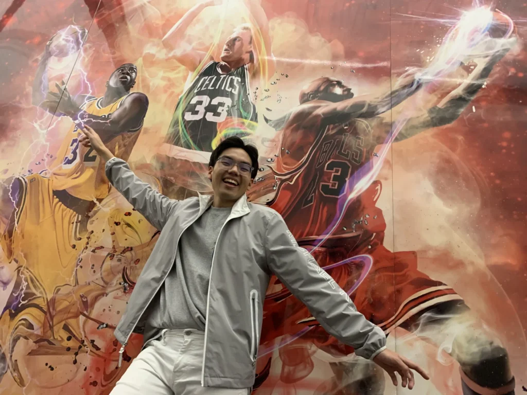A person poses energetically, arms extended, in front of a dynamic mural featuring basketball players in vibrant action. Players wear jerseys with numbers 32, 33, and 23, amidst swirling colors.
